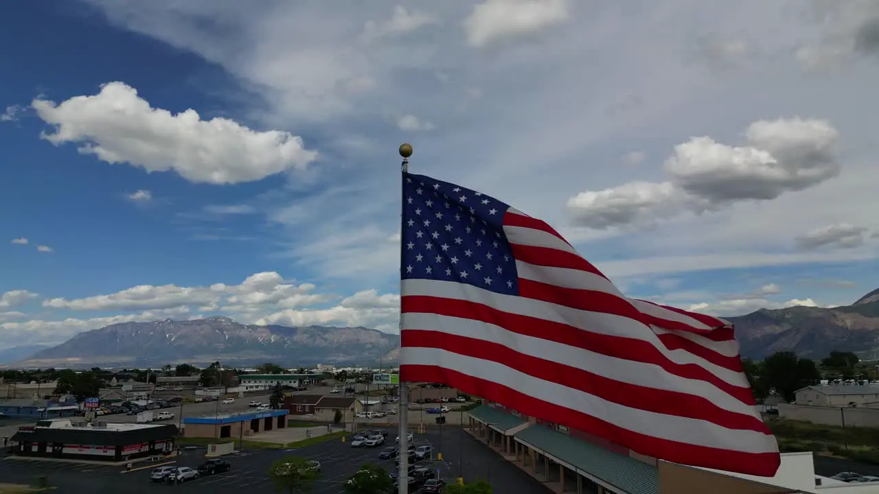 American flag USA blowing waving in the wind on beautiful sunny summer day with clouds and blue skies overlooking small town America USA and mountains as drone flys pans around flagpole in 4K 60fps