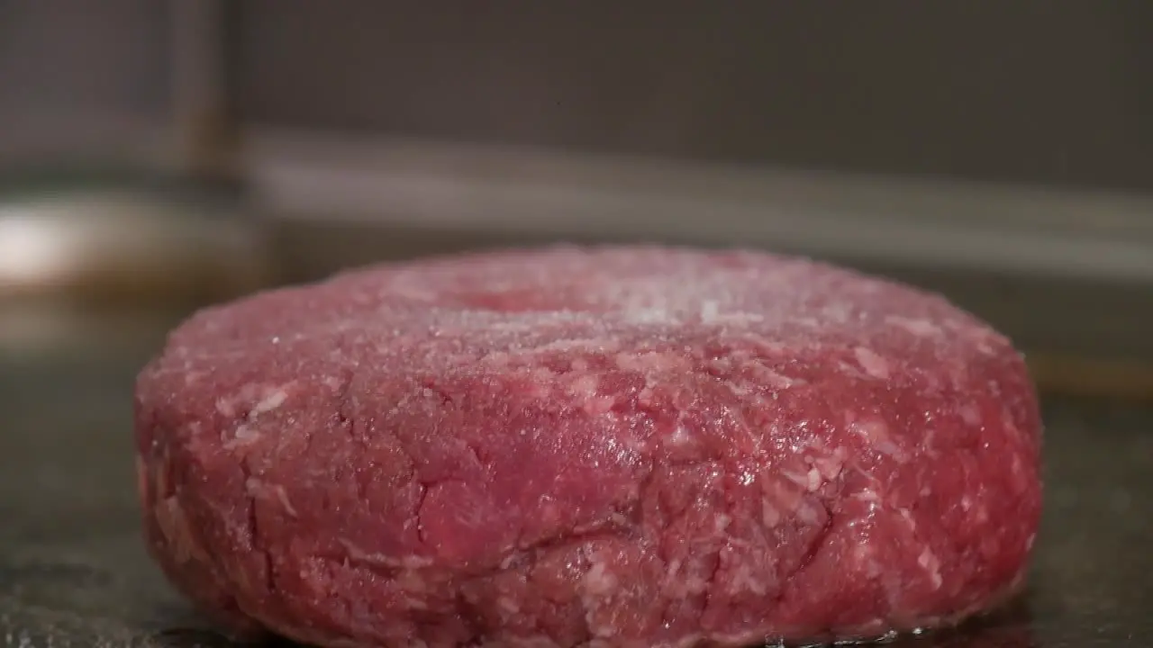 Close-up of a raw burger patty on a restaurant grill as black pepper and salt are ground on top