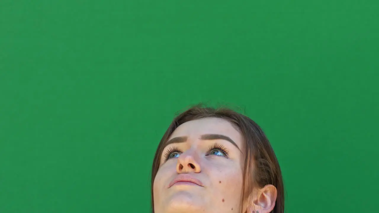 Static close-up shot of a brunette lady's face from the nose up in front of a green chroma key backdrop looking at the sky with excitement and laughter