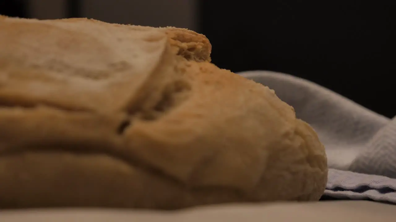 Slow motion pan into freshly baked loaf of sour dough bread topped with flour sitting on kitchen bench with tea towel and tray low depth of field