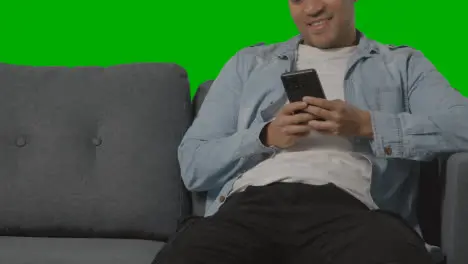 Close Up Of Young Man Sitting On Sofa Looking At Mobile Phone Against Green Screen