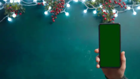 Overhead Shot Of Person Holding Green Screen Mobile Phone With Christmas Decorations And Holly Berries 1