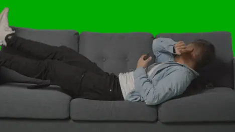 Young Man Lying On Sofa Sneezing And Looking At Mobile Phone Against Green Screen