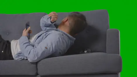 Studio Shot Of Man Lying On Sofa Scrolling Through Mobile Phone And Sneezing Against Green Screen