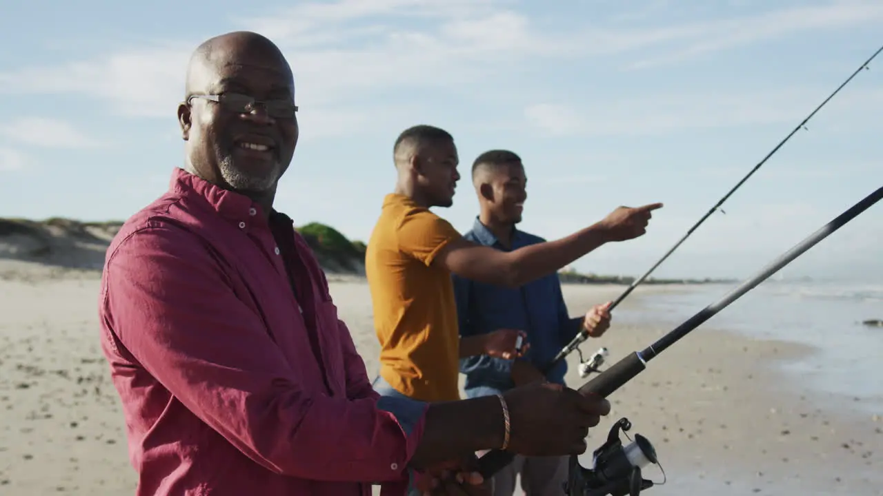 African american senior father and twin teenage sons standing on a beach fishing and talking