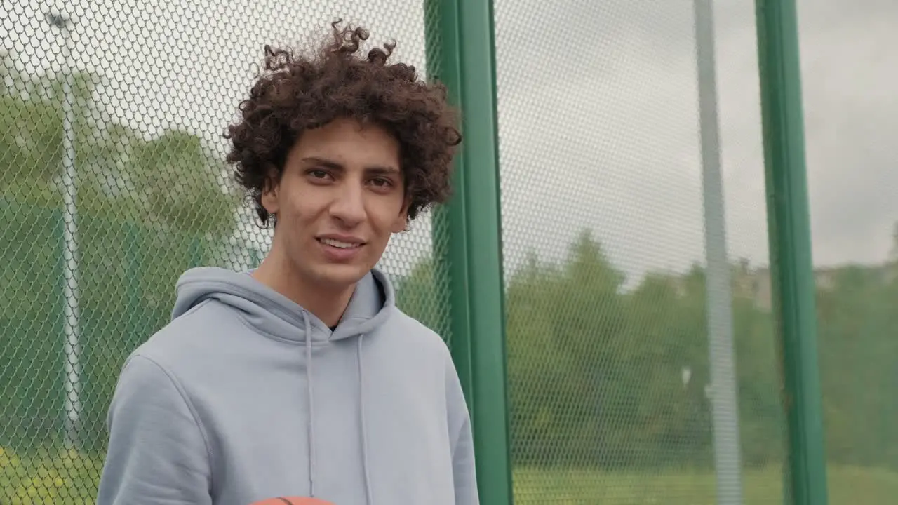 Young Smiling Active Male Basket Player In Gray Sweatshirt Holding Ball While Standing On Playground