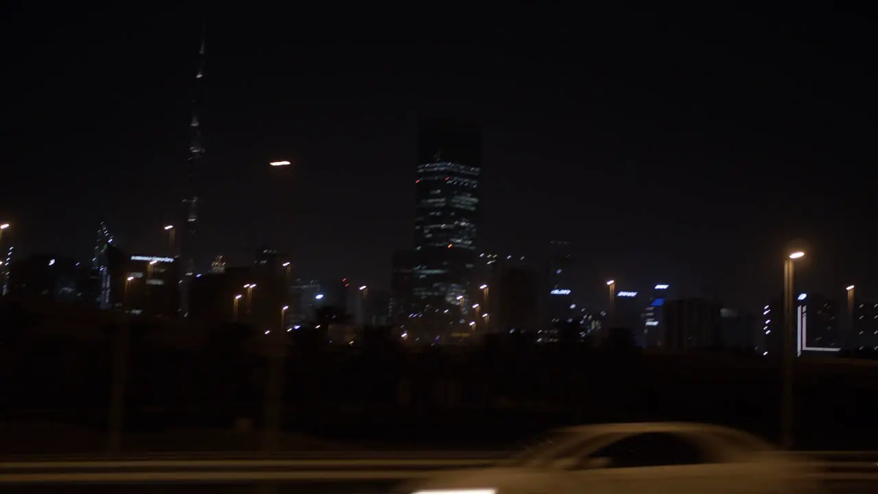 Driving in a car and passing by Dubai skyline at night in a car on the highway with distant lit up buildings and skyscrapers including the Burj Khalifa in the United Arab Emirates