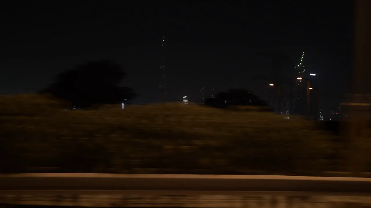 Passing by Dubai skyline at night in a car on the highway with distant lit up buildings and skyscrapers including the Burj Khalifa in the United Arab Emirates