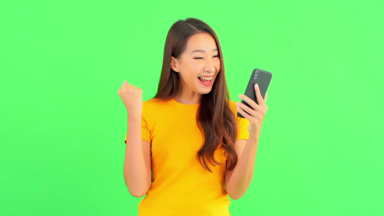 Young brunette woman using her mobile phone isolated on a green screen then surprised checking something and starts to celebrate as if she won a lottery