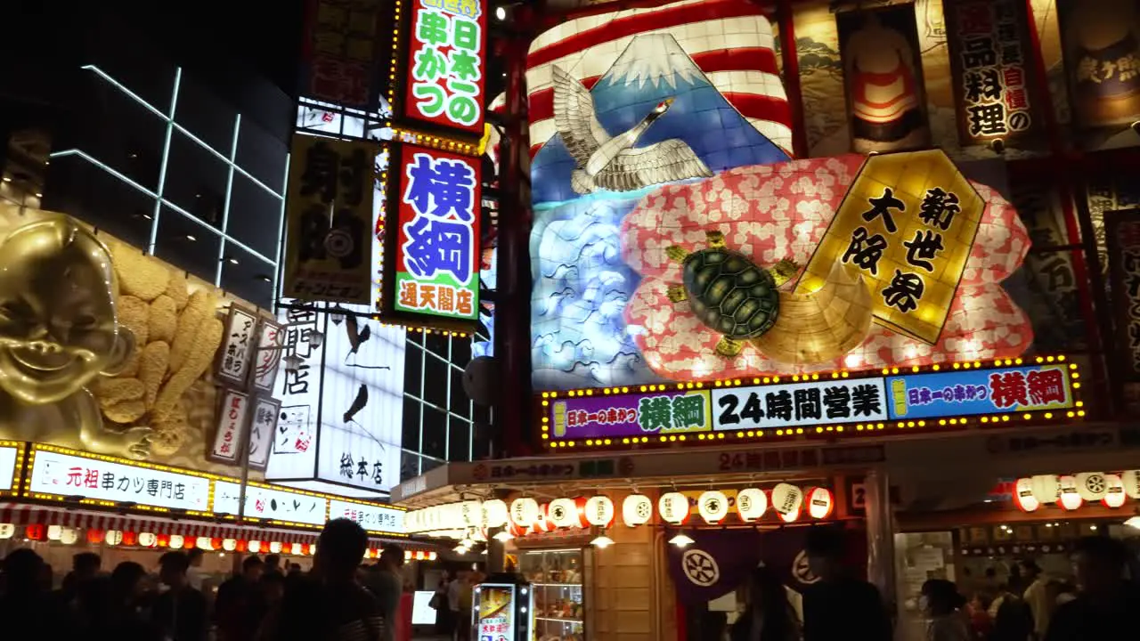 Pan Right Shot Across Illuminated Colourful Display Above Restaurant With Biliken Statue On Corner In Shinsekai Area At Night