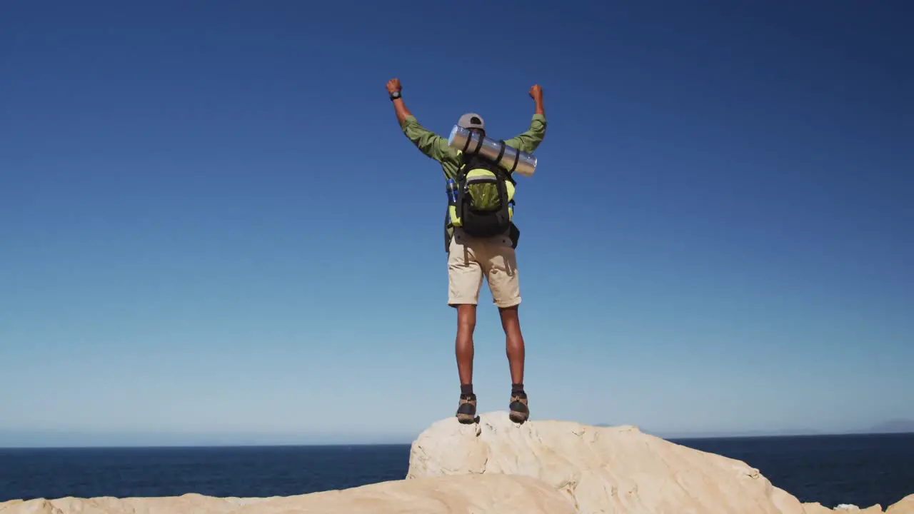 African american man hiking in mountains standing on rock raising his hands by the sea