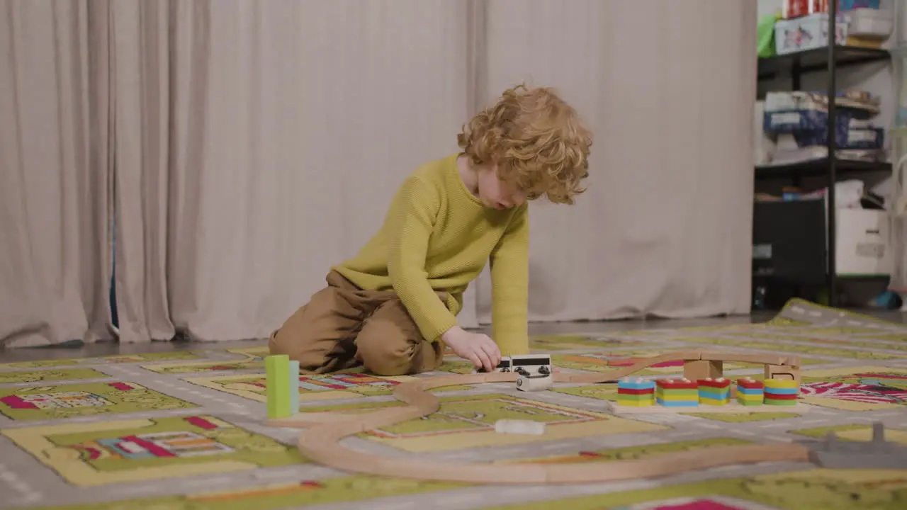 Little Blond Boy Playing With Wooden Cars On The Carpet In Classroom In A Montessori School