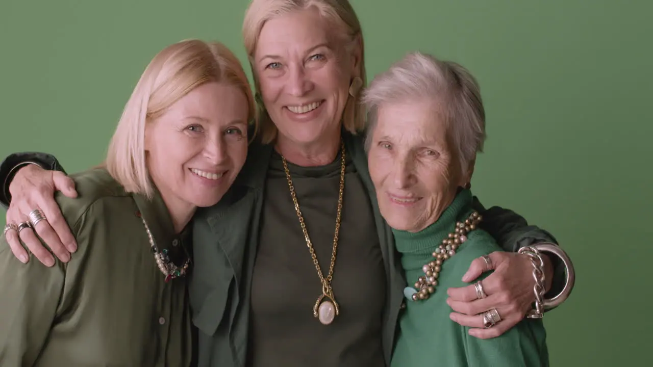 Two Blonde Mature Women And Blonde Senior Woman Smiling And Hugging Wearing Green Tones Clothes And Posing On Green Background