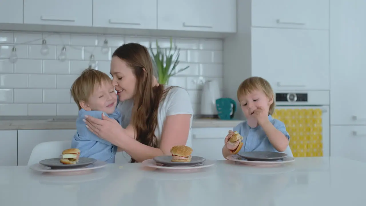 A young beautiful mother in a white dress with two children is smiling and eating fresh burgers in her kitchen Happy family homemade food healthy foods