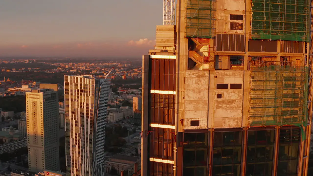 Fly around top of skyscraper construction site high above city Concrete walls and scaffolding lit by setting sun Warsaw Poland