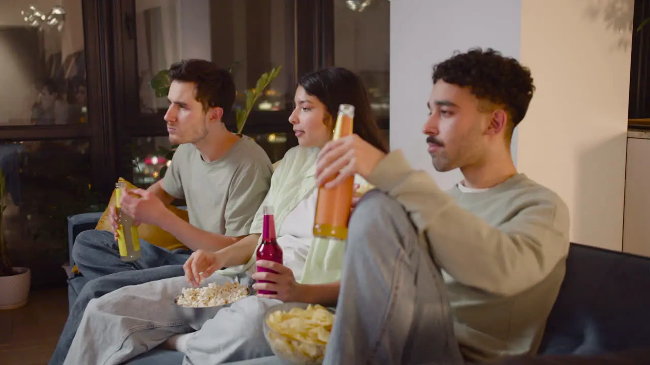 Vista Lateral De Tres Amigos Viendo Una Película Interesante En La Televisión Sentados En El Sofá Comiendo Palomitas De Maíz Y Papas Fritas Y Bebiendo Refrescos 1