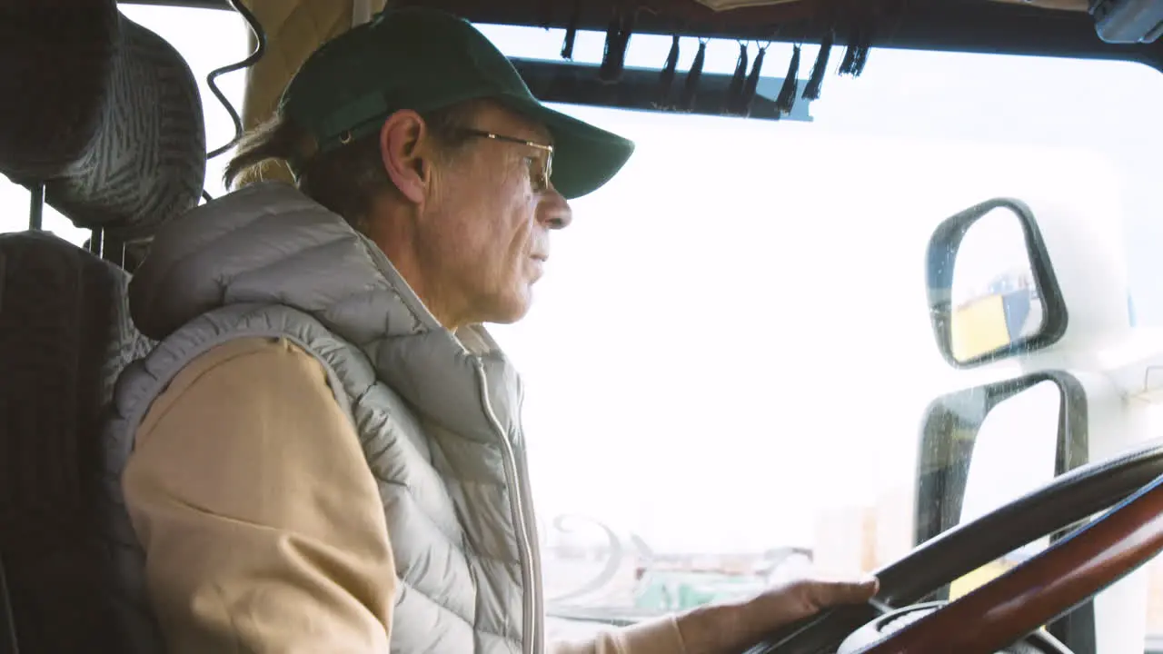 Close Up View Of Older Worker Wearing Cap And Vest Driving A Truck In A Logistics Park