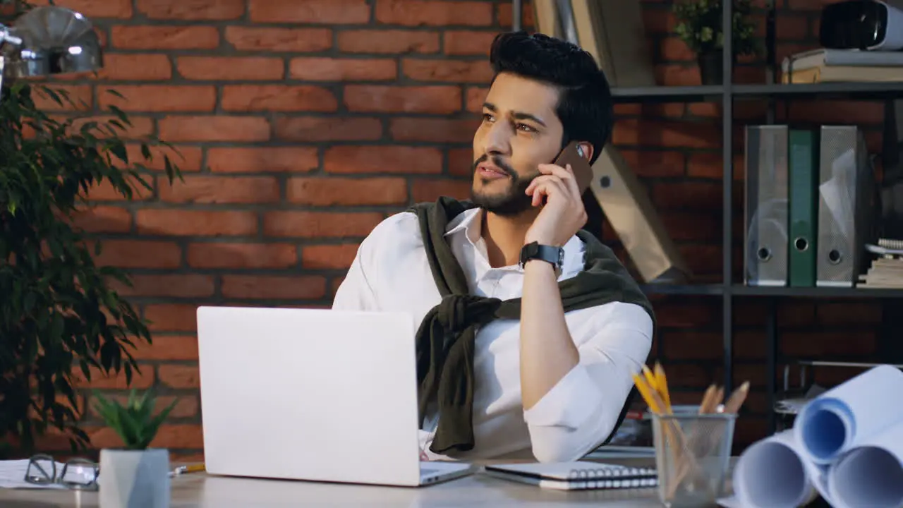 Young Arabian Businessman Sitting At The Laptop Computer Sitting At Desk And Cheerfully Talking On The Phone