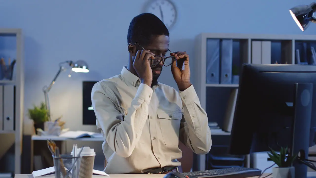 Office Worker Sitting At Desk With Computer Trying To Rest While Massaging His Neck And Taking Off His Glasses In The Office At Night