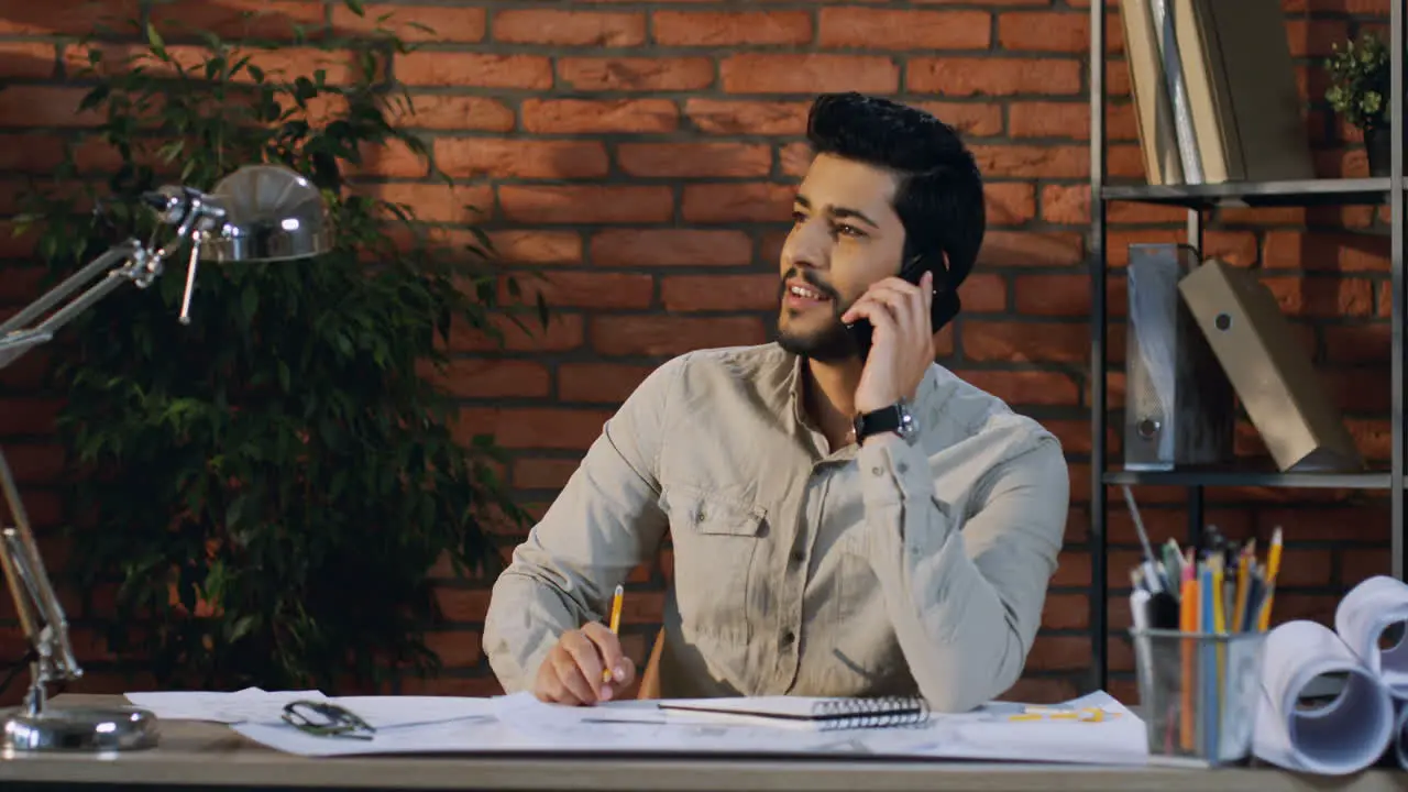 Young Man Architect Talking On The Phone Cheerfully While Sitting At The Desk With Drawing On It