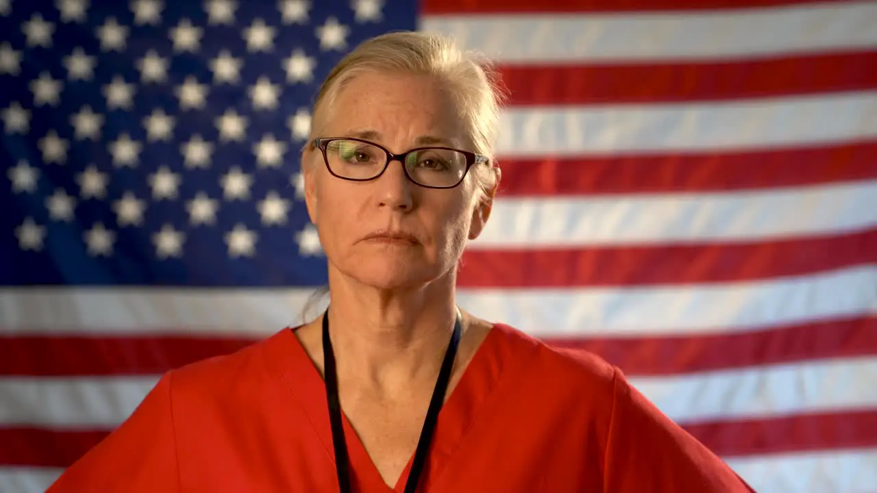 Medium tight portrait of a healthcare nurse looking concerned and sympathetic walking towards the camera with an out of focus American flag