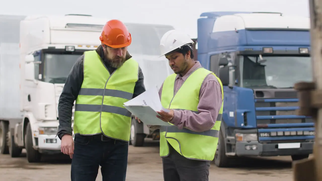 Boss And Worker Wearing Vests And Safety Helmets Organizing A Truck Fleet In A Logistics Park While They Consulting A Document 2