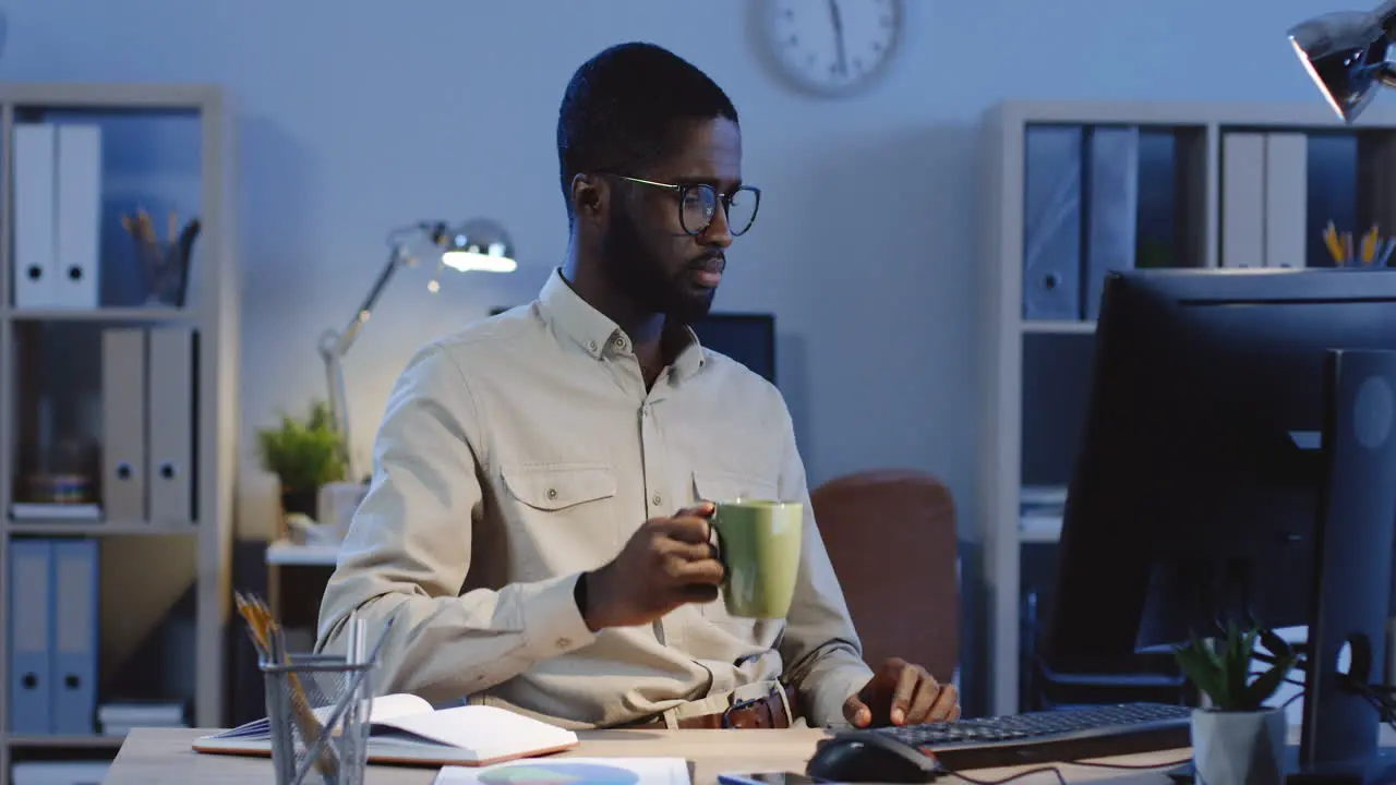 Young Man Drinking Coffee While Working At The Computer In The Office At Night