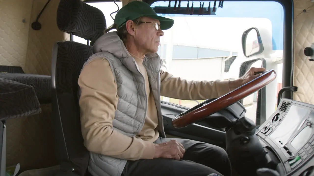 Side View Of Older Worker Wearing Cap And Vest Driving A Truck In A Logistics Park 1