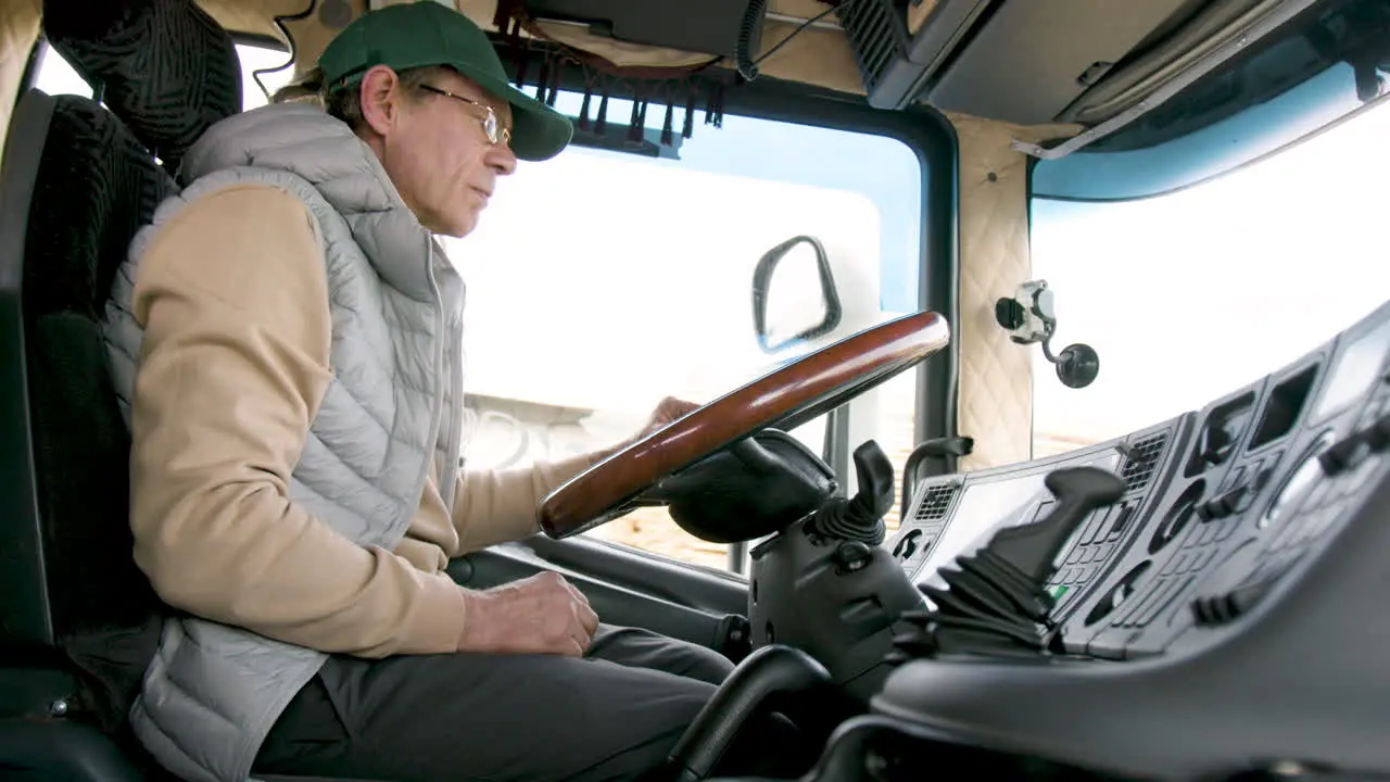 Side View Of Older Worker Wearing Cap And Vest Driving A Truck In A Logistics Park 4