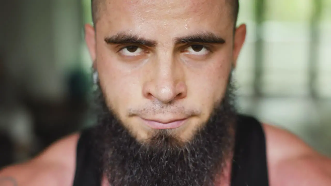 Young man with beard looks into the camera sweating and breathing strong after exercise in gym close up