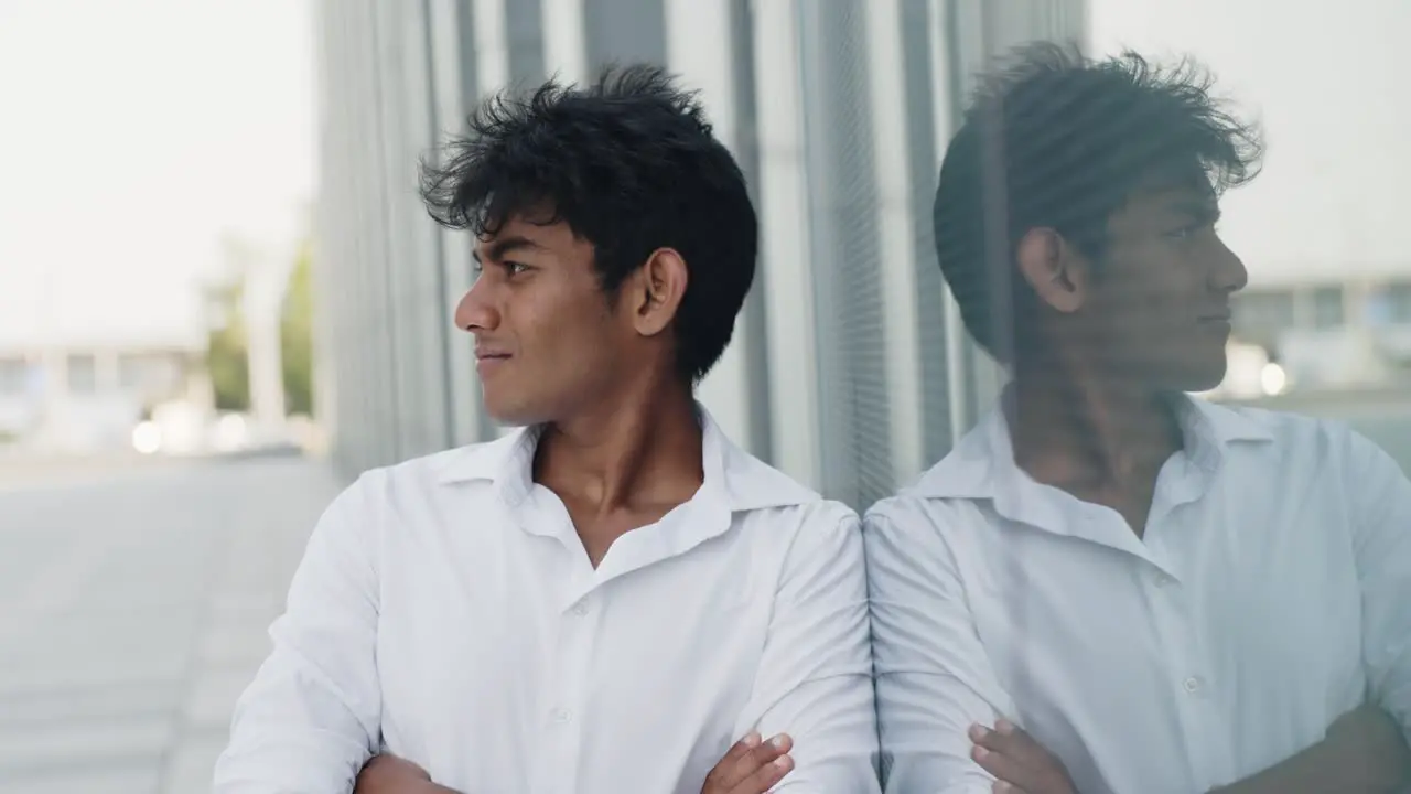 Indian young guy crossing his arms leaning against a glass wall on the street