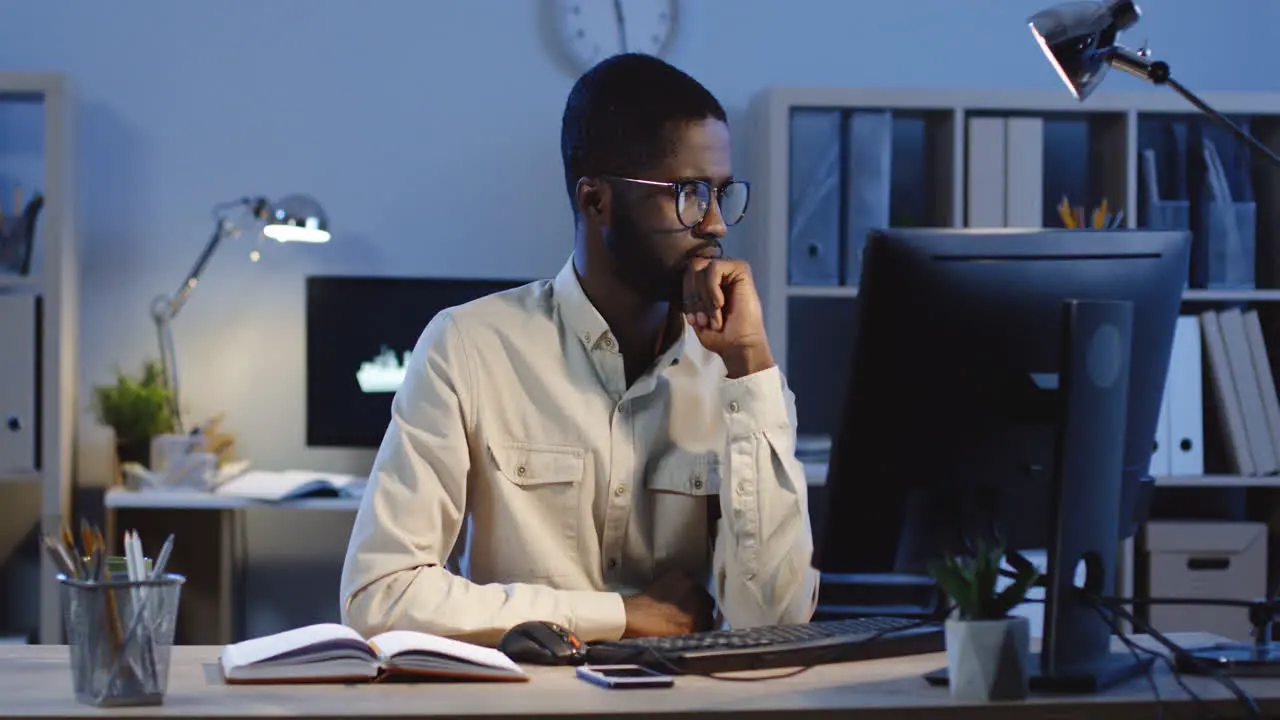 Office Worker In Glasses Thinking Seriously In The Office Looking At The Screen Of Computer And Making Notes In His Notebook In The Office At Night