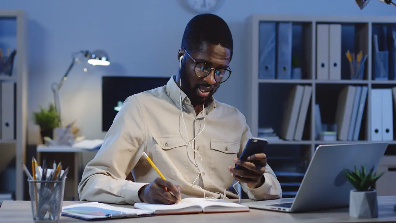 Young Office Worker In Headphones Sitting At The Computer Using His Smartphone And Writing In The Notebook While Looking At The Screen