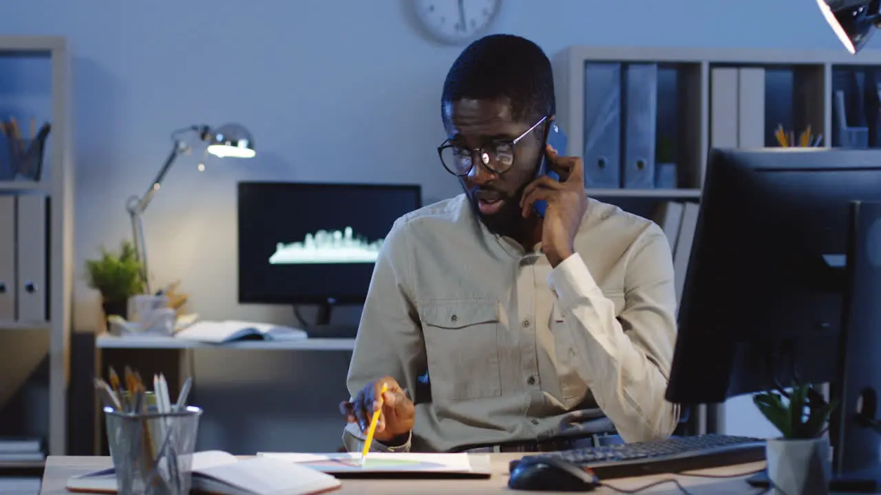 Young Worried Businessman Talking On The Phone And Consulting About Some Documents Of Project Sitting At Desk In The Office At Night