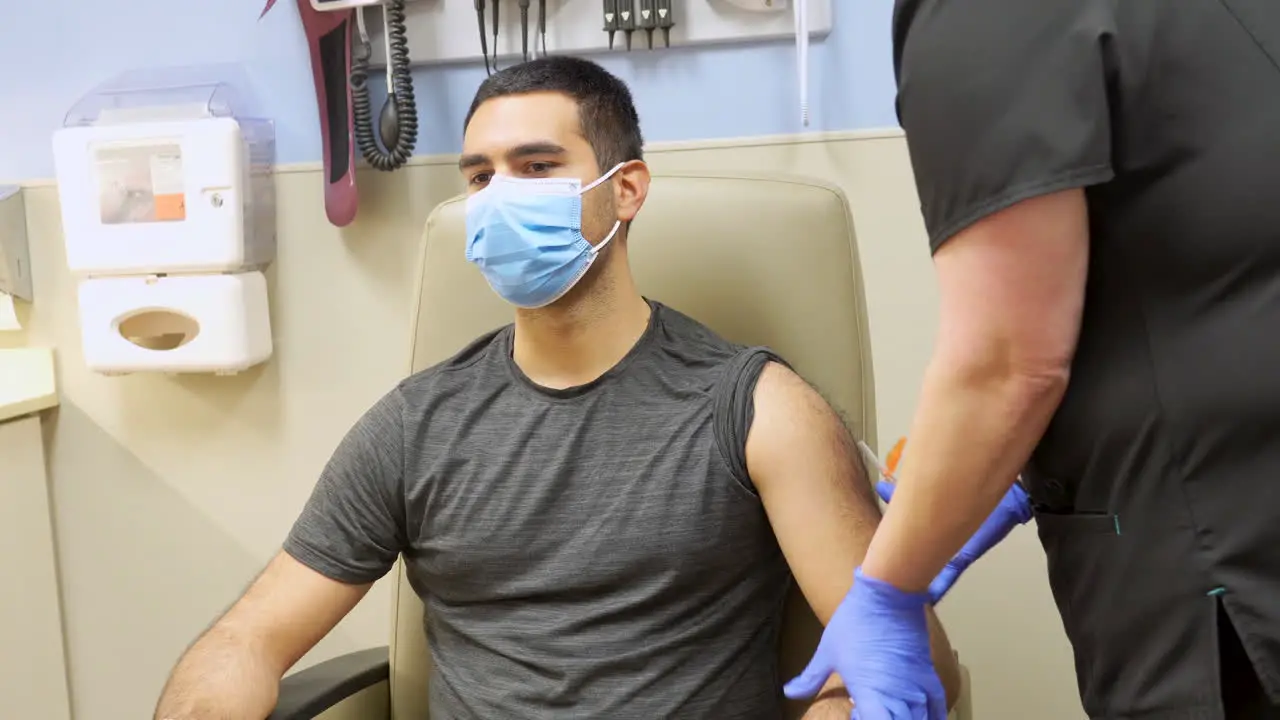 Nurse Cleans Shoulder and Administers COVID 19 Vaccine Shot in the Arm of Young Adult Dark Skinned Man of Minority Ethnicity Wearing a Medical Mask Corona Virus Injection in Doctor’s Office