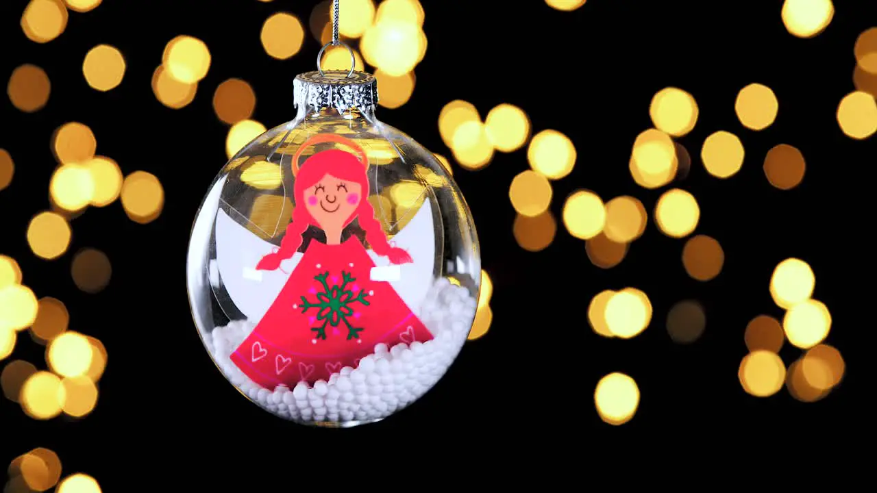 single clear Christmas ball ornament with cartoon red hair angel and snow inside out of focus lights flickering in the background close up