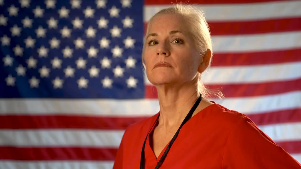 Medium tight portrait of the back of blonde nurses head as she turns and looks at camera looking angry and concerned with out of focus US flag