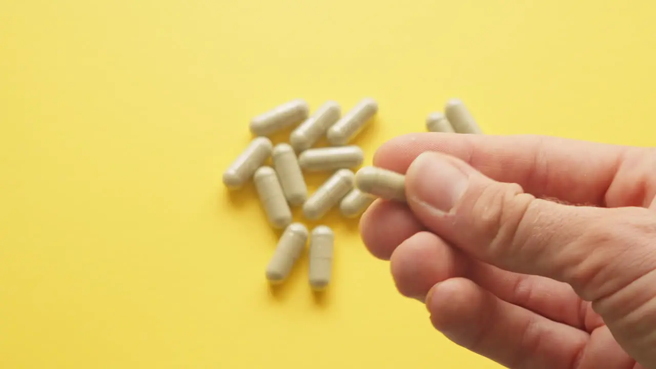 Right Male Hand Holding Lions Mane Supplement Pill In Between Fingers With Yellow Background With Pills On Table