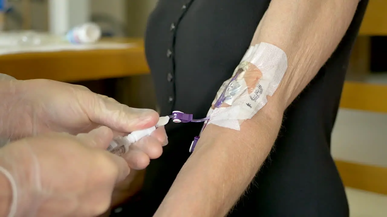 Medical nurse doing an at home iv patient visit wearing sterile gloves while finishing a saline solution injection from a syringe into a picc line and capping it off