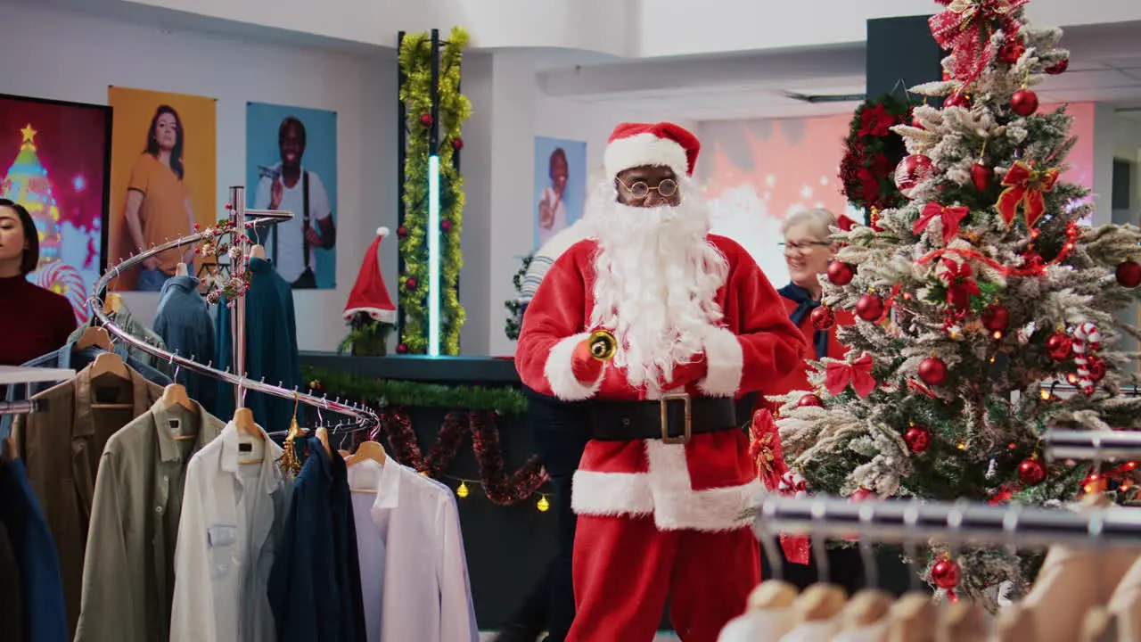 Dolly in shot of merry manager dressed as Santa Claus in Christmas adorn shopping mall clothing shop Supervisor jingling xmas bells and greeting customers in holiday themed fashion store