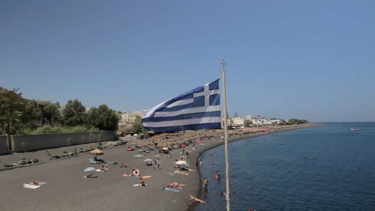 Wide shot of the Kamari beach in Santorini with a Greek flag flapping in the foreground