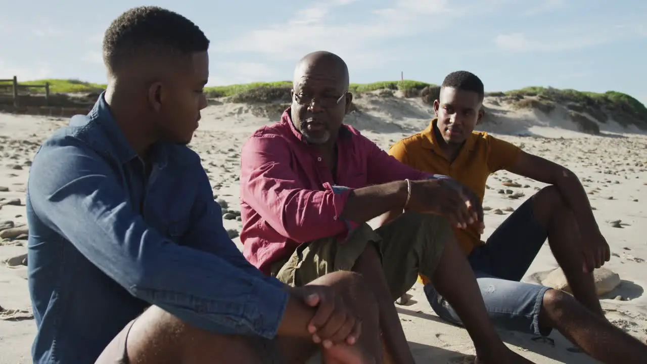 African american senior father and twin teenage sons sitting on a beach talking