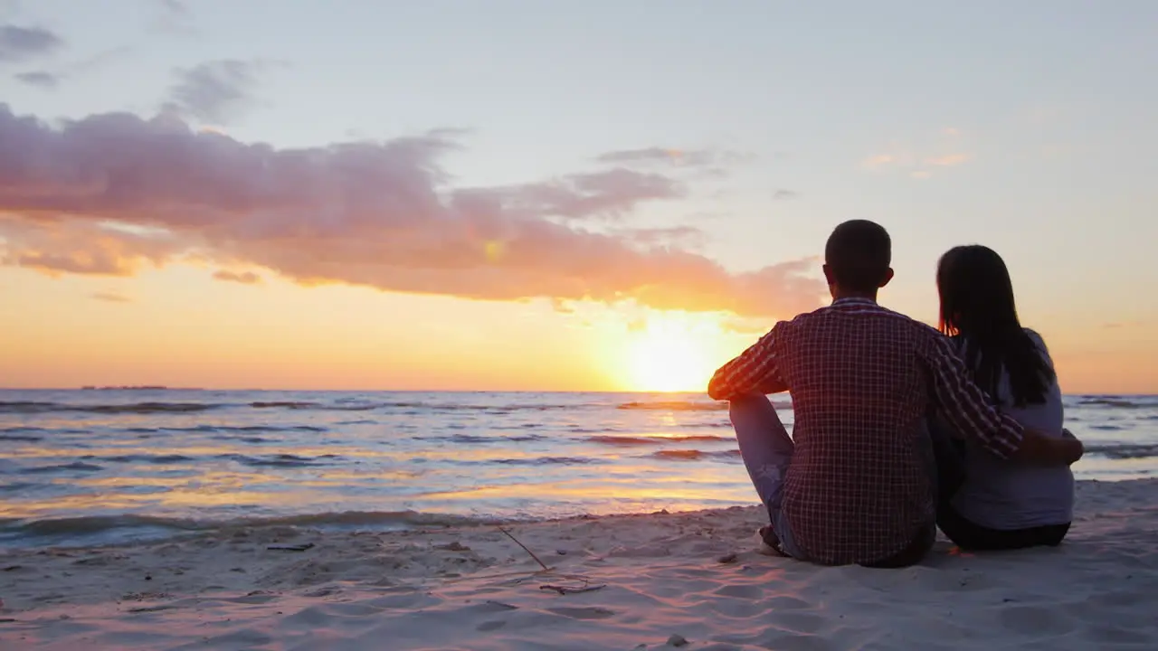 Young Romantic Couple Sitting On The Beach Admiring The Sunset