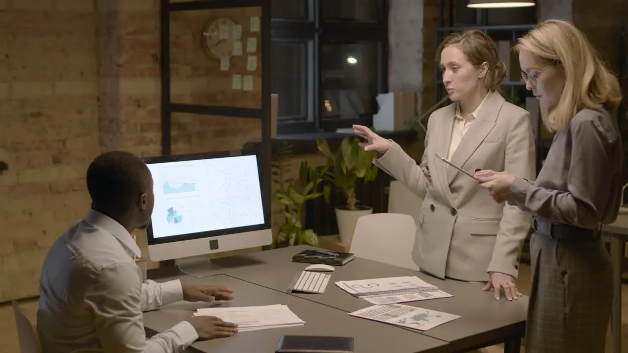 American Man Sitting At Desk In The Office While Talking About Graphics And Stadistics With Two Female Coworkers Who Are Standing