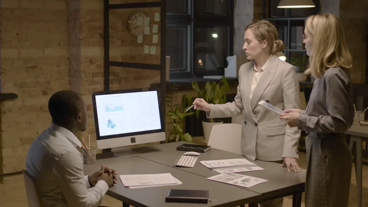 American Man Sitting At Desk In The Office While Talking About Graphics And Stadistics With Two Female Coworkers Who Are Standing 1