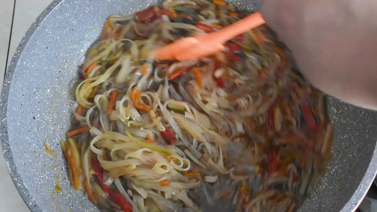 Man's hand stirring noodles and chopped vegetables while frying in a wok