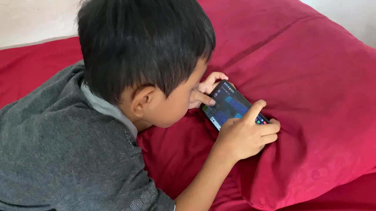 A 4 year old asian boy playing games on his smartphone while lying in bed