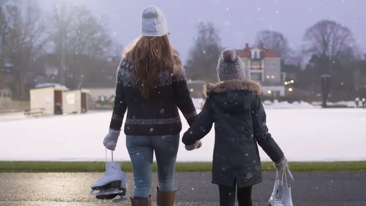 Middle aged mom and 8 year old young daughter walks hand in hand together on a snowy winters day to the ice skating rink to practice ice skating for bonding activities as a family during Christmas fun