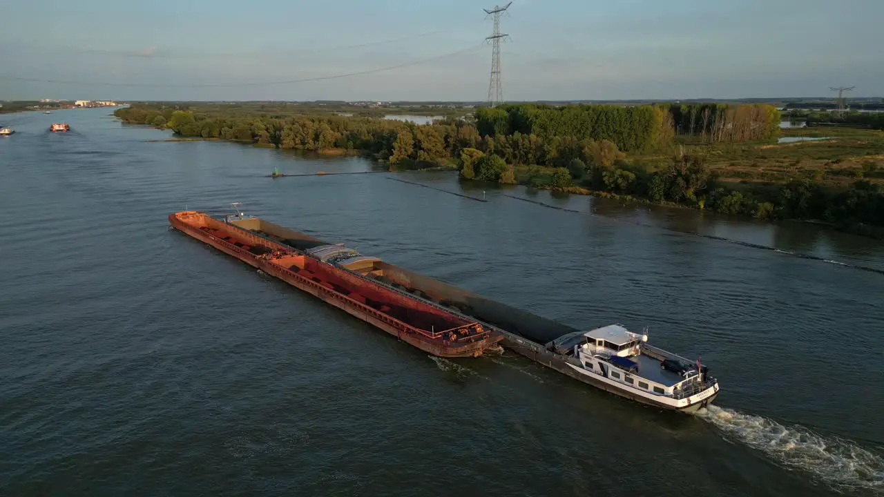 Aerial slide and pan footage of long cargo ship transporting bulk material in barges on river in flat landscape