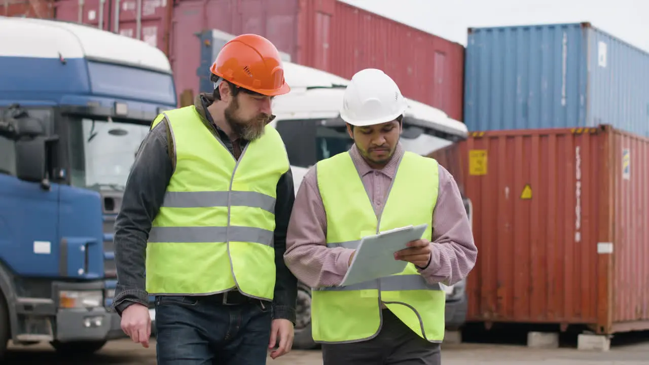 Boss And Worker Wearing Vests And Safety Helmets Organizing A Truck Fleet In A Logistics Park While They Consulting A Document And Walking